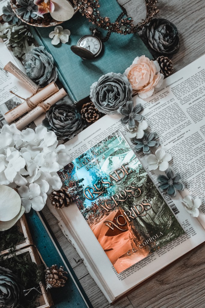 Flatlay photo of Crusade Across Worlds by C.G. Coppola. Surrounded by books, flowers, pinecones, and a pocket watch, the fantasy cover blends icy blue mountains with fiery desert landscapes, symbolizing interworld conflict.